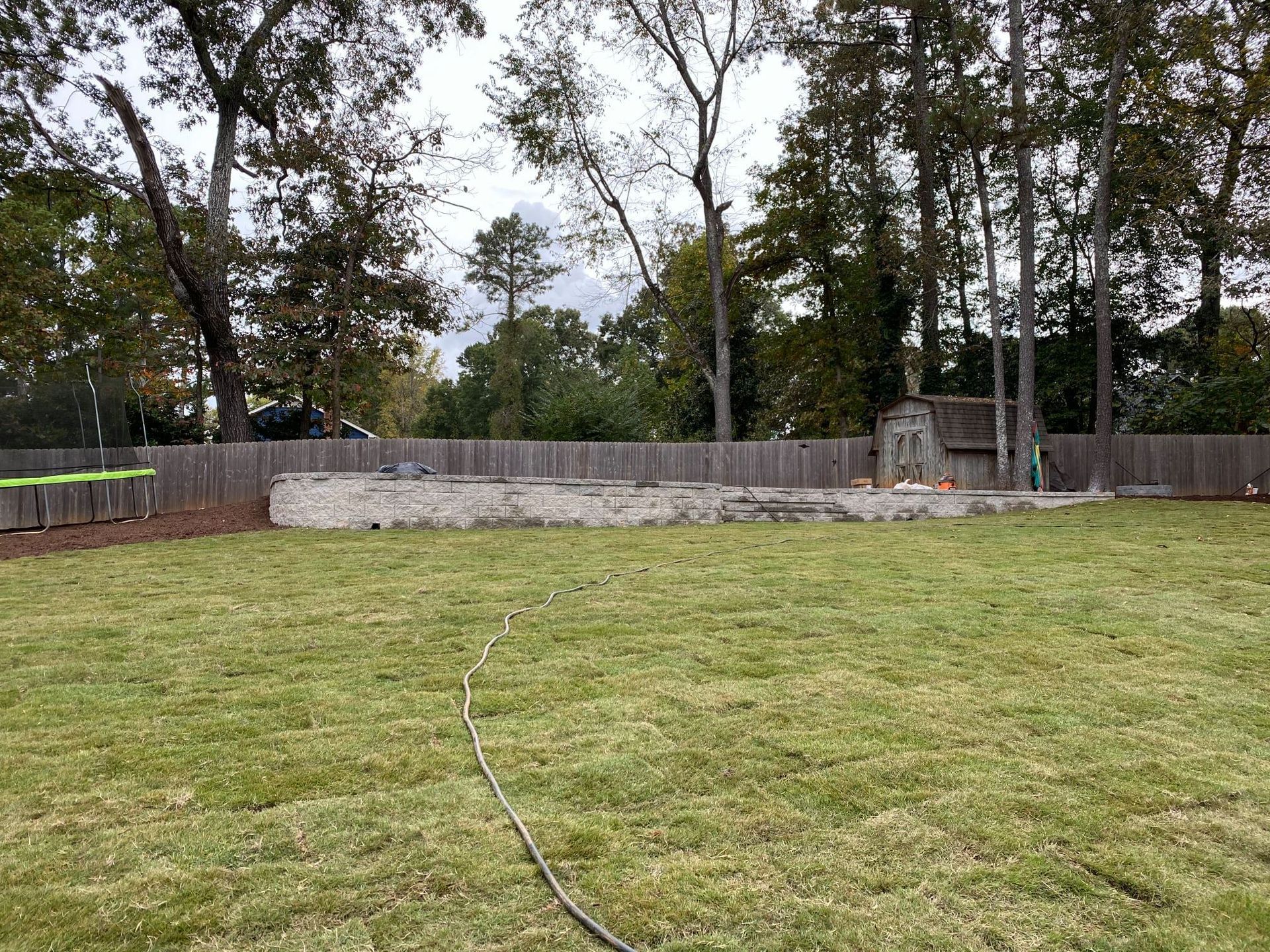 Grassy backyard with a stone retaining wall, wooden fence, and trees in the background