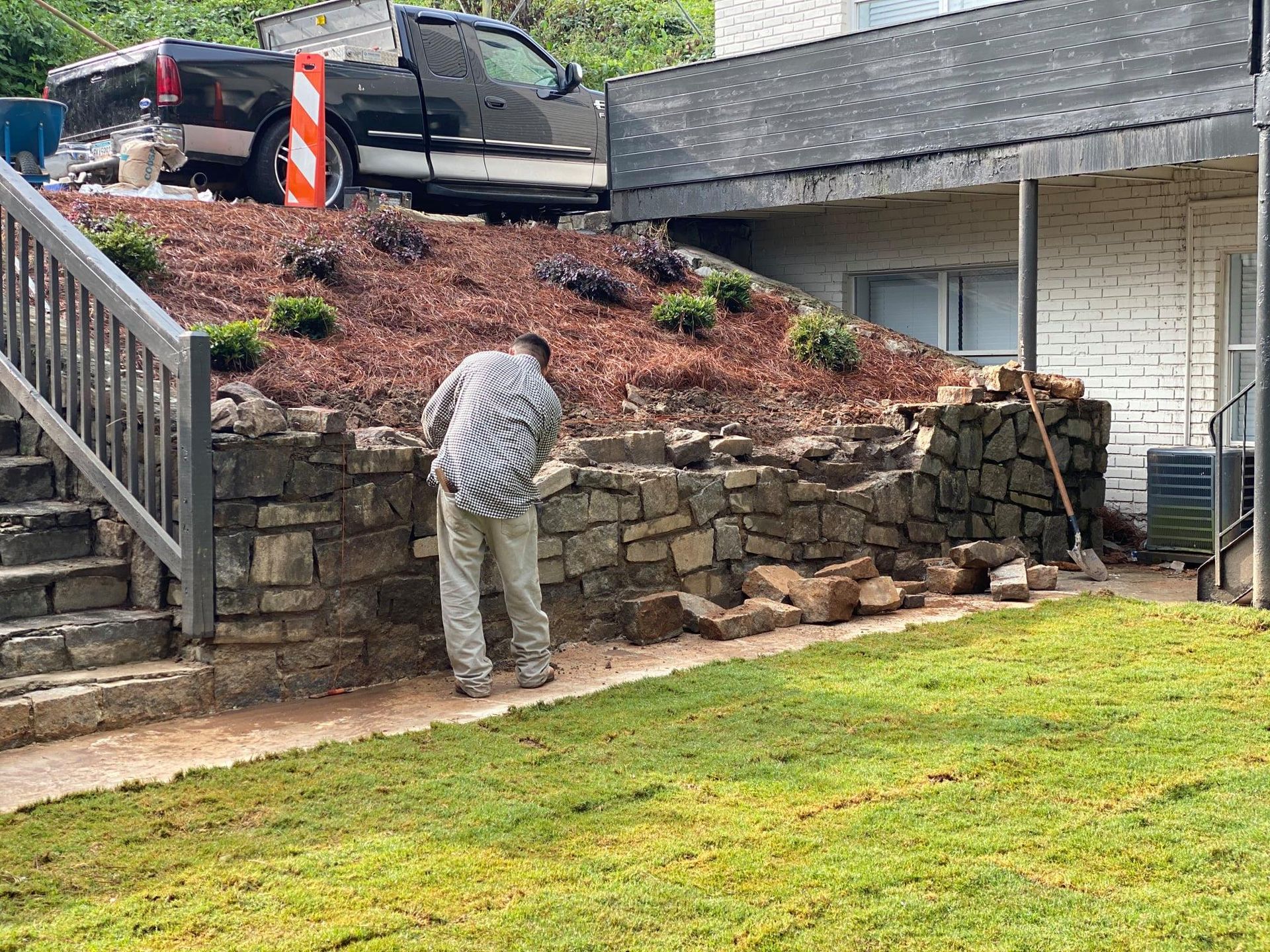 Person working on a stone retaining wall in a front yard beside stairs and a parked pickup truck.