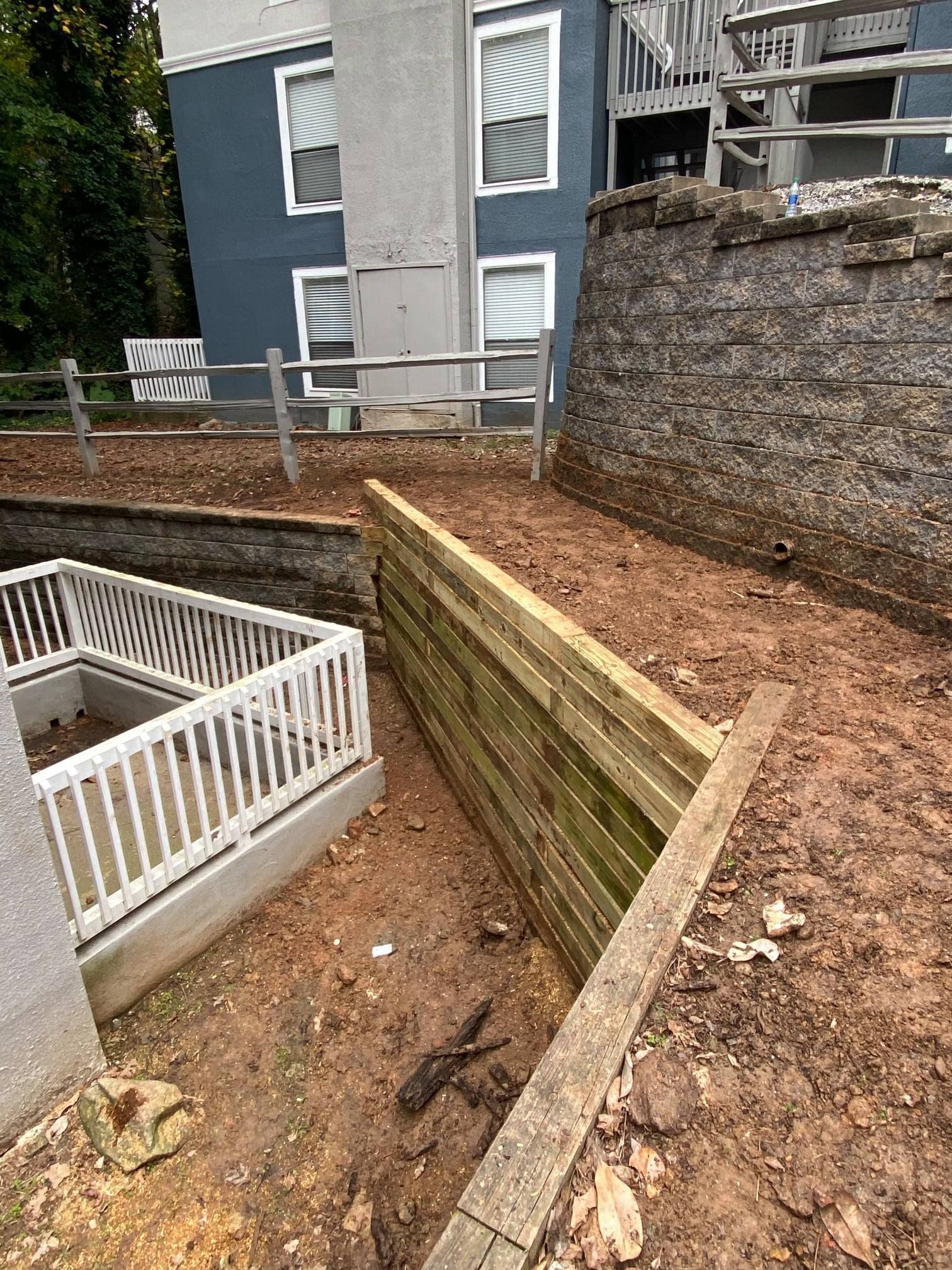 Wooden retaining wall beside an outdoor stairwell and dirt slope next to a house