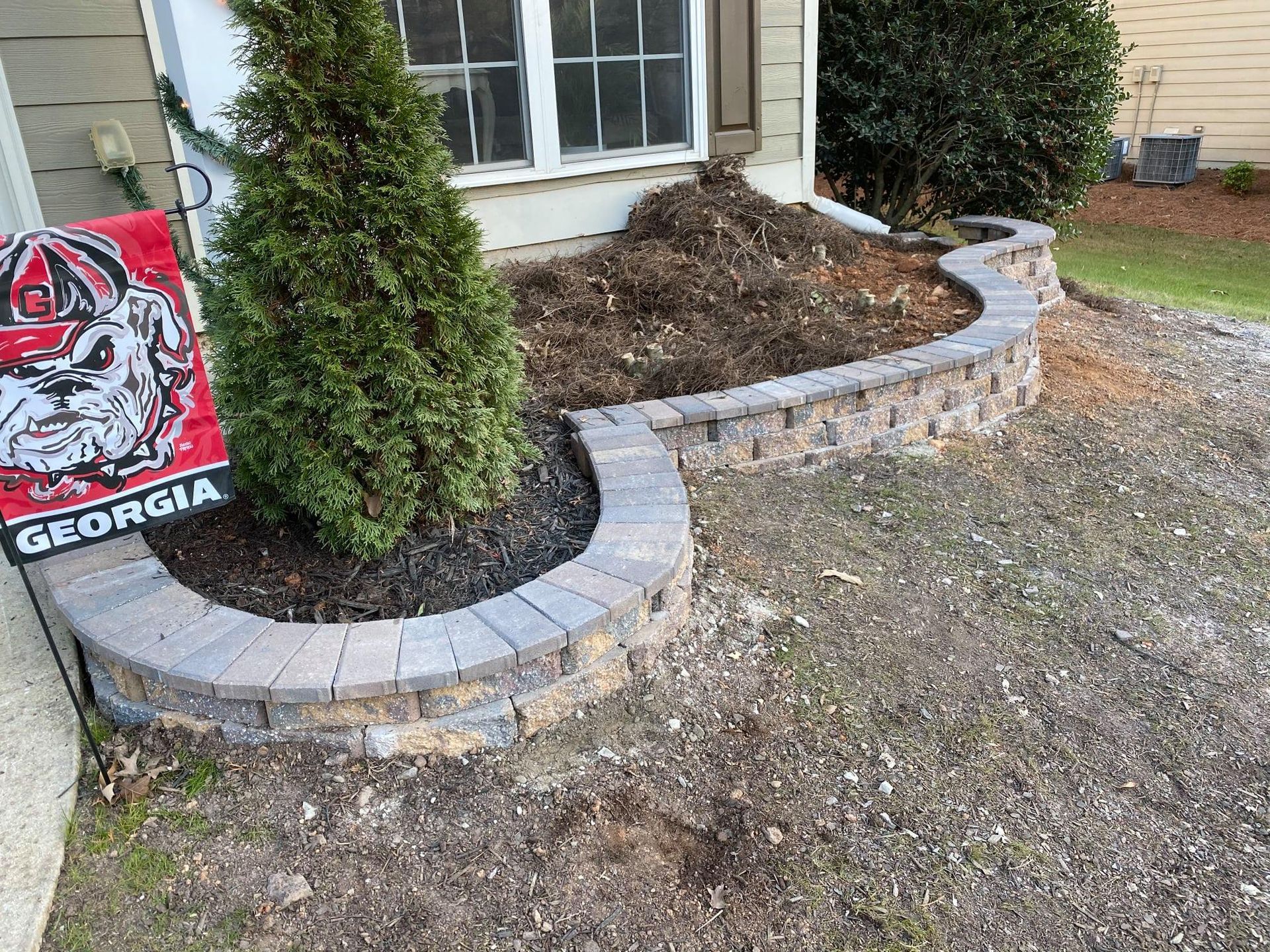 Curved stone garden bed with shrubs beside a house window and a Georgia flag on the left