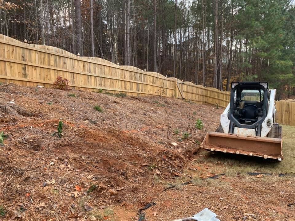 Compact skid steer grading a dirt slope beside a wooden fence in a wooded yard.