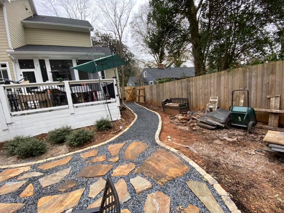 Curving stone walkway beside a house, with a wooden fence and outdoor equipment along the yard.