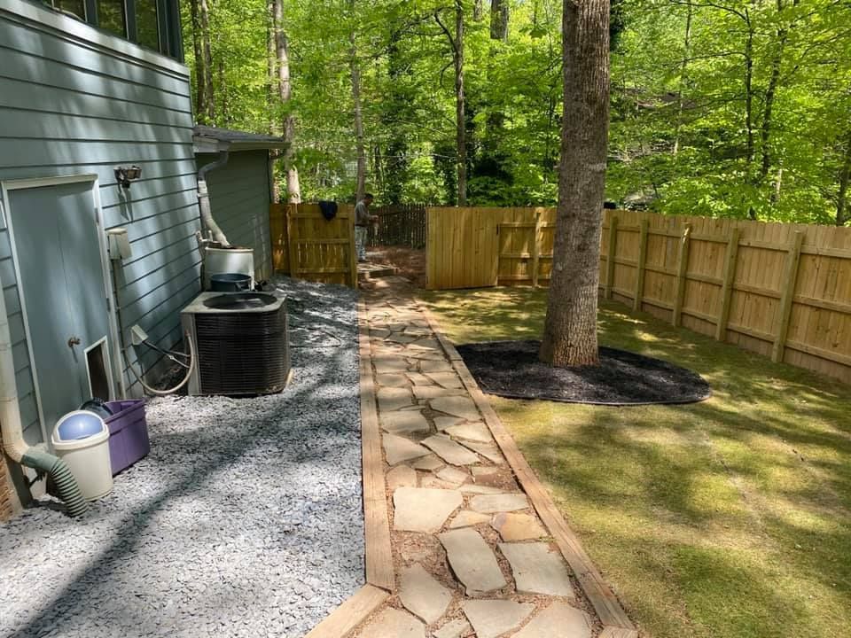 Backyard path beside gray house, gravel area, tree, and wooden fence in a wooded yard