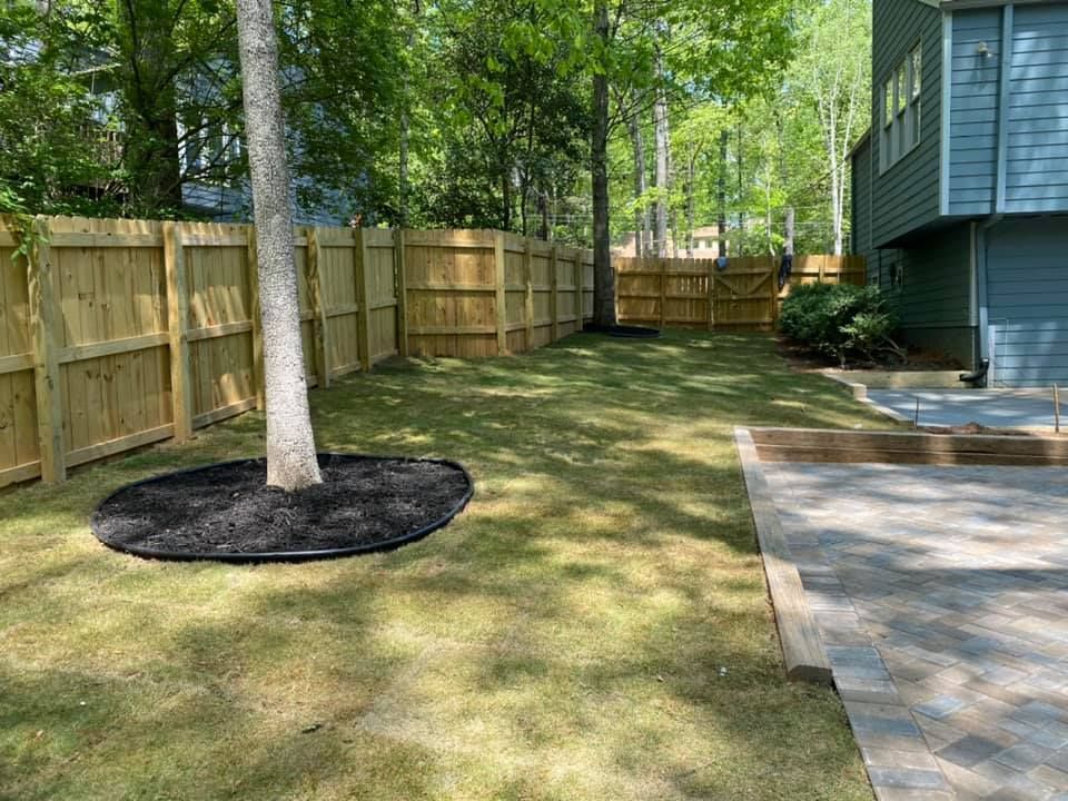Backyard with wooden fence, trees, grass, and a house patio.