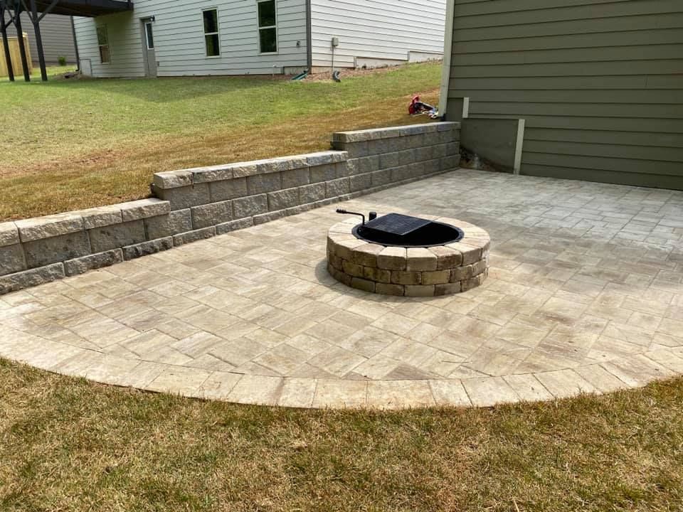 Patio with circular stone fire pit, tan pavers, and low retaining wall beside a house and lawn