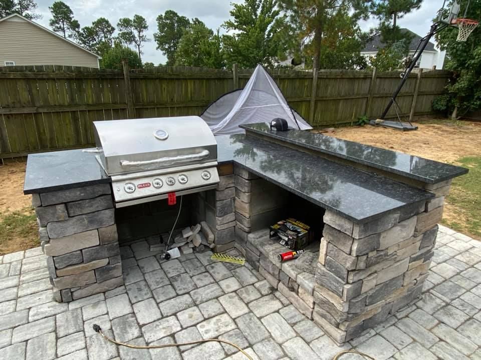 Patio with circular stone fire pit, tan pavers, and low retaining wall beside a house and lawn