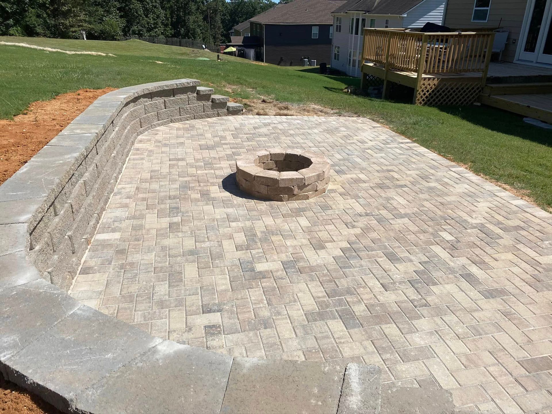 Paver patio with a circular stone fire pit and curved retaining wall beside a grassy yard