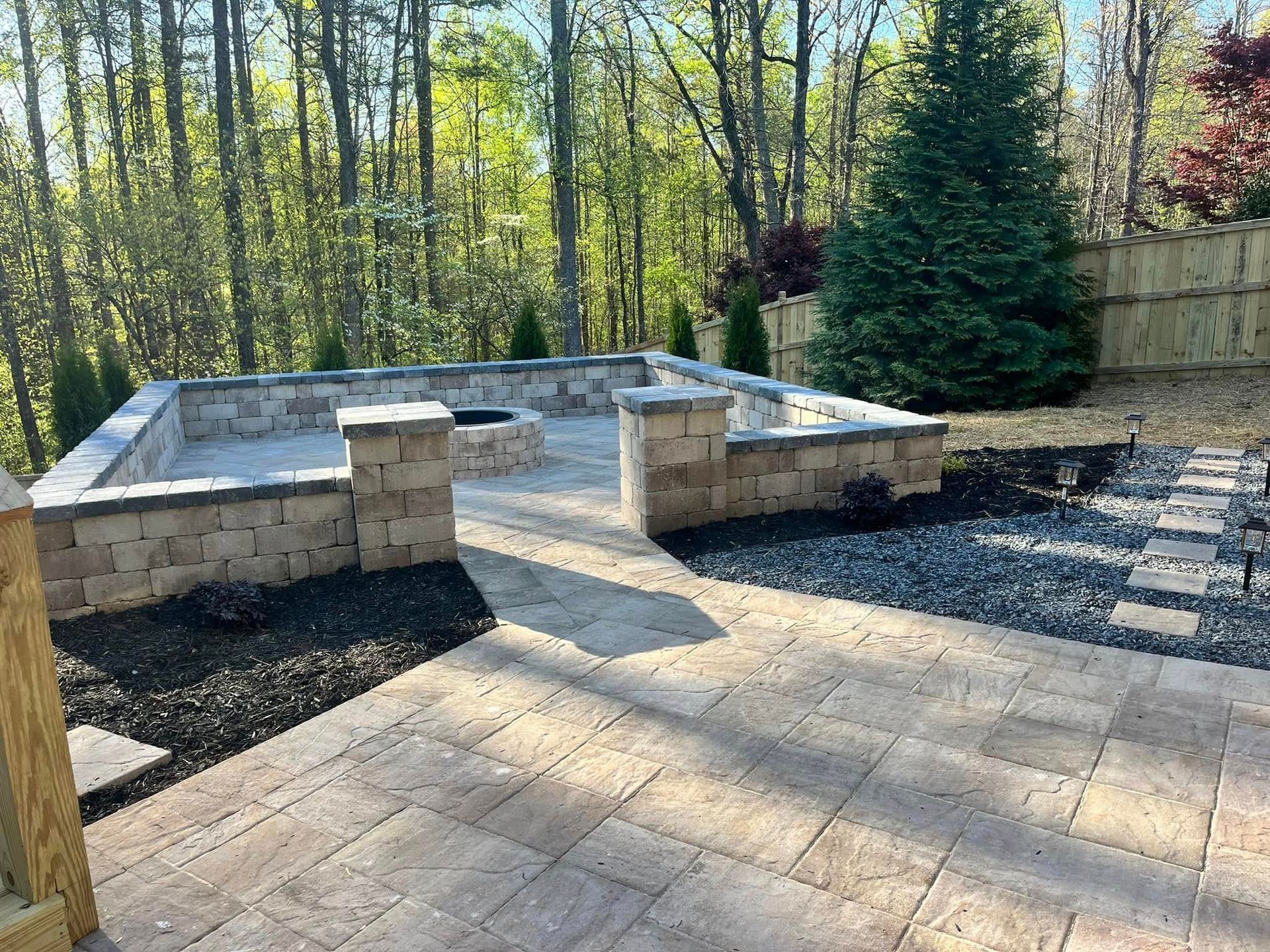 Backyard patio with stone pavers and raised foundation walls beside trees and a privacy fence