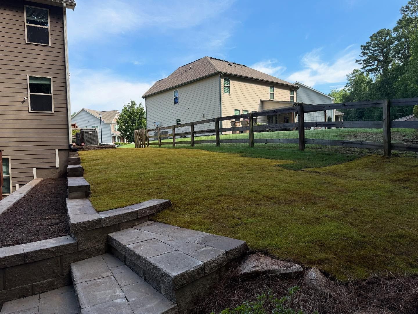 Backyard with stone steps, grassy slope, black fence, and a two-story house under a blue sky