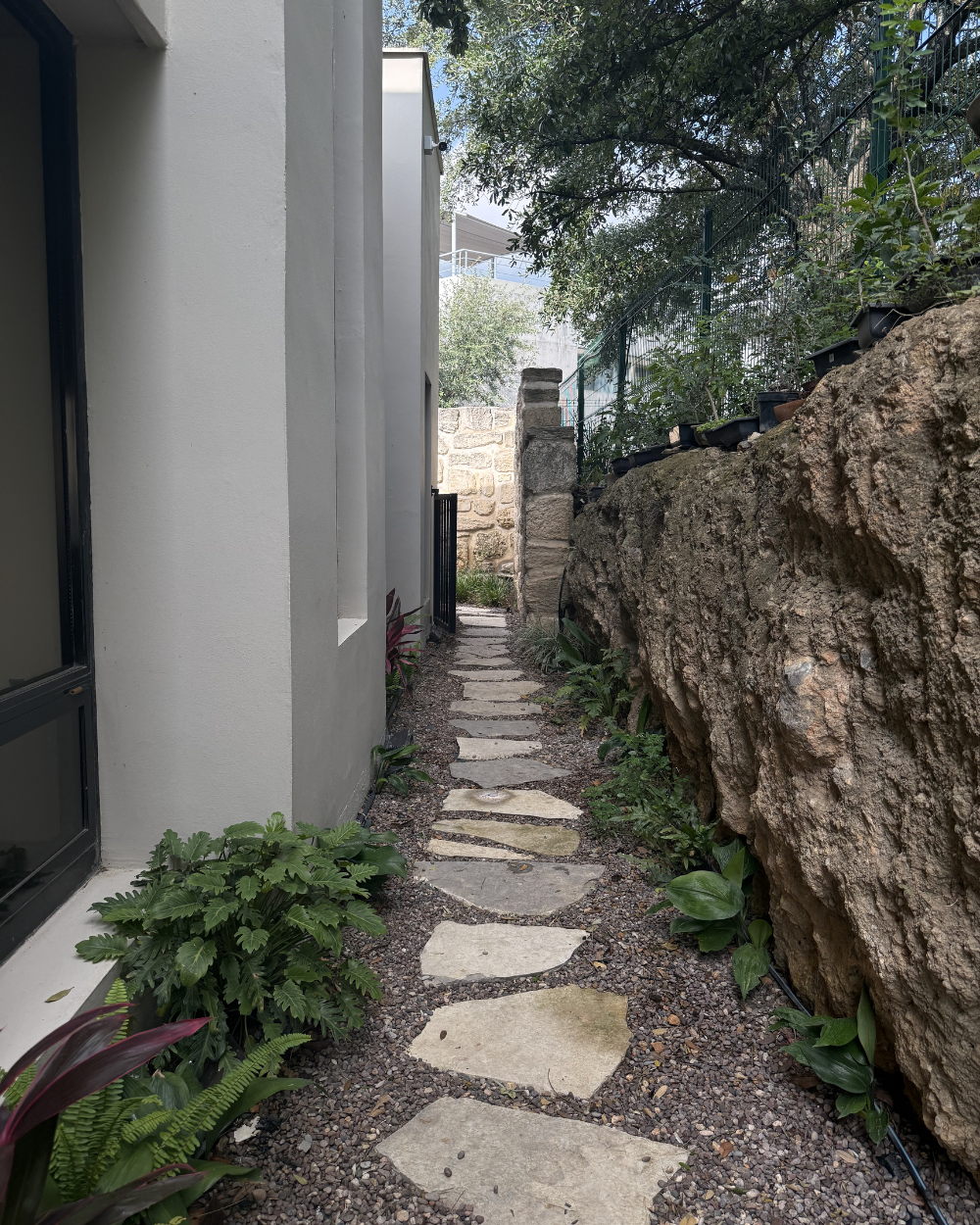 Un estrecho sendero de piedra junto a una casa y un muro de piedra, que atraviesa un frondoso corredor ajardinado.
