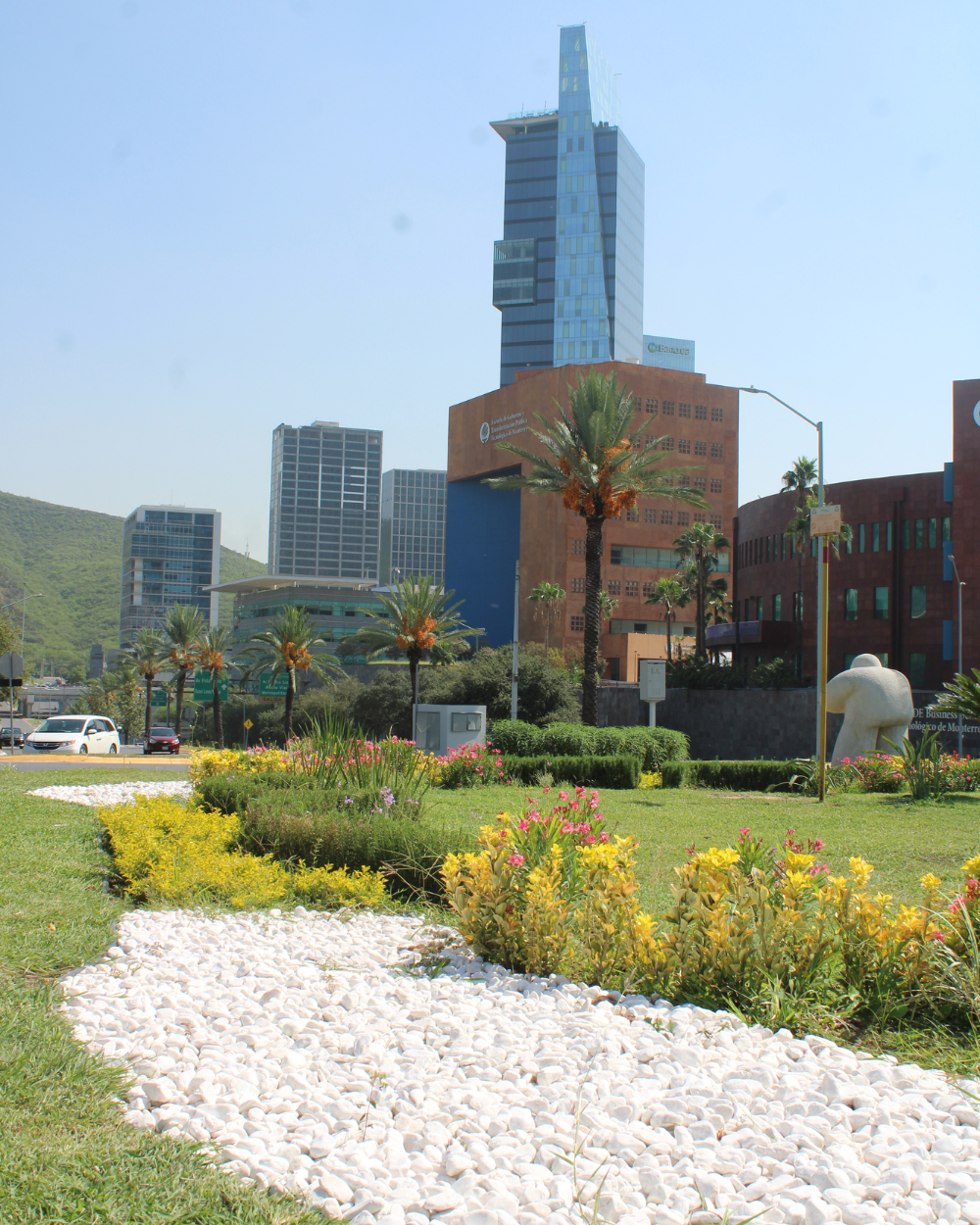 Parque urbano con parterres de flores y sendero de grava blanca, con altos rascacielos modernos al fondo.
