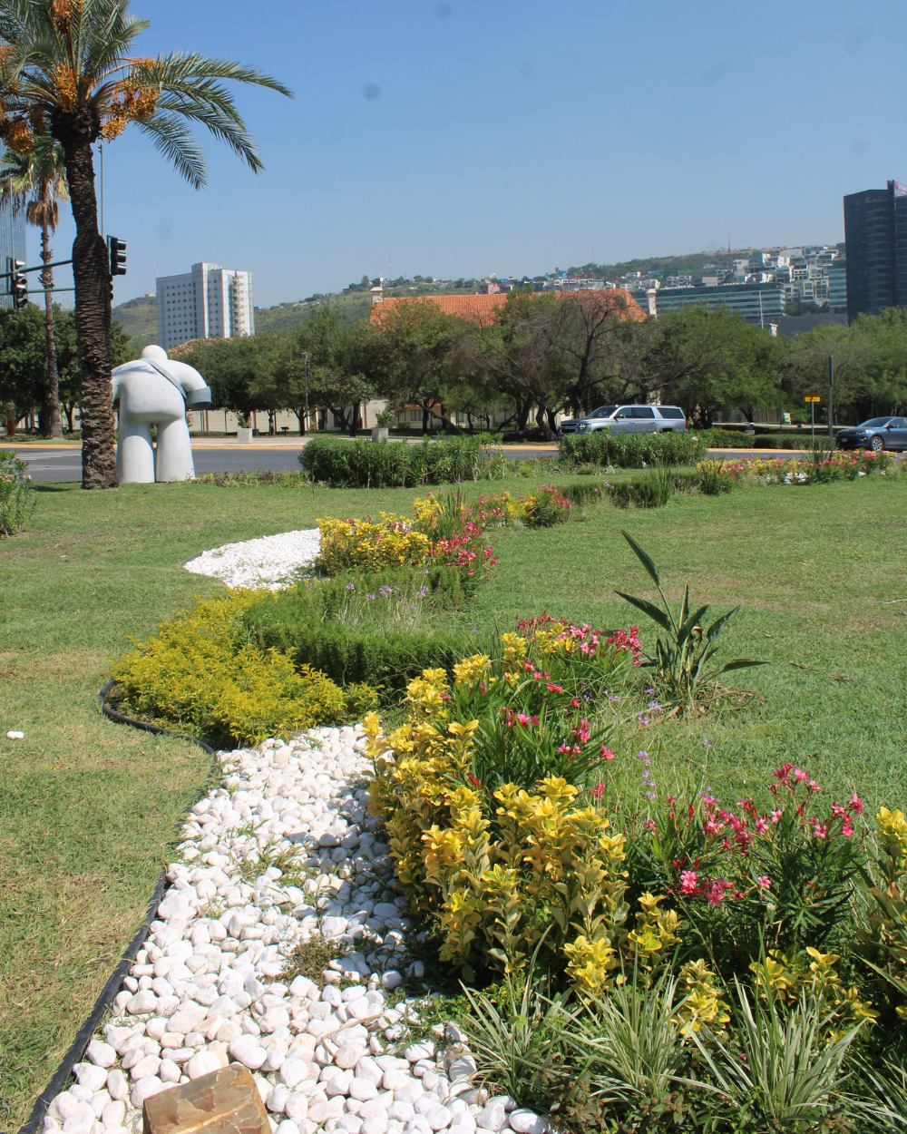 Jardín paisajístico con un borde sinuoso de piedra blanca, flores coloridas, palmeras y edificios a lo lejos bajo un cielo azul.