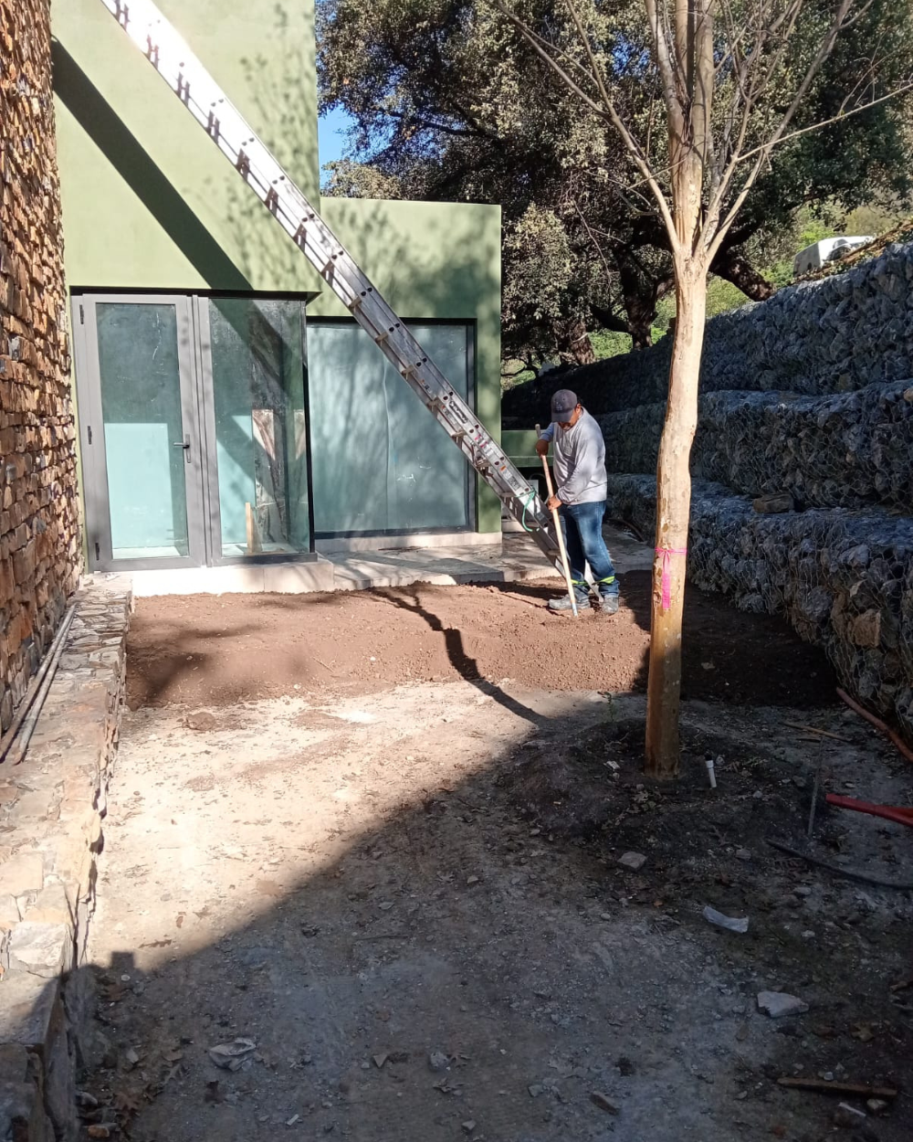 Persona pintando de verde una pared exterior junto a una puerta en un camino de tierra.