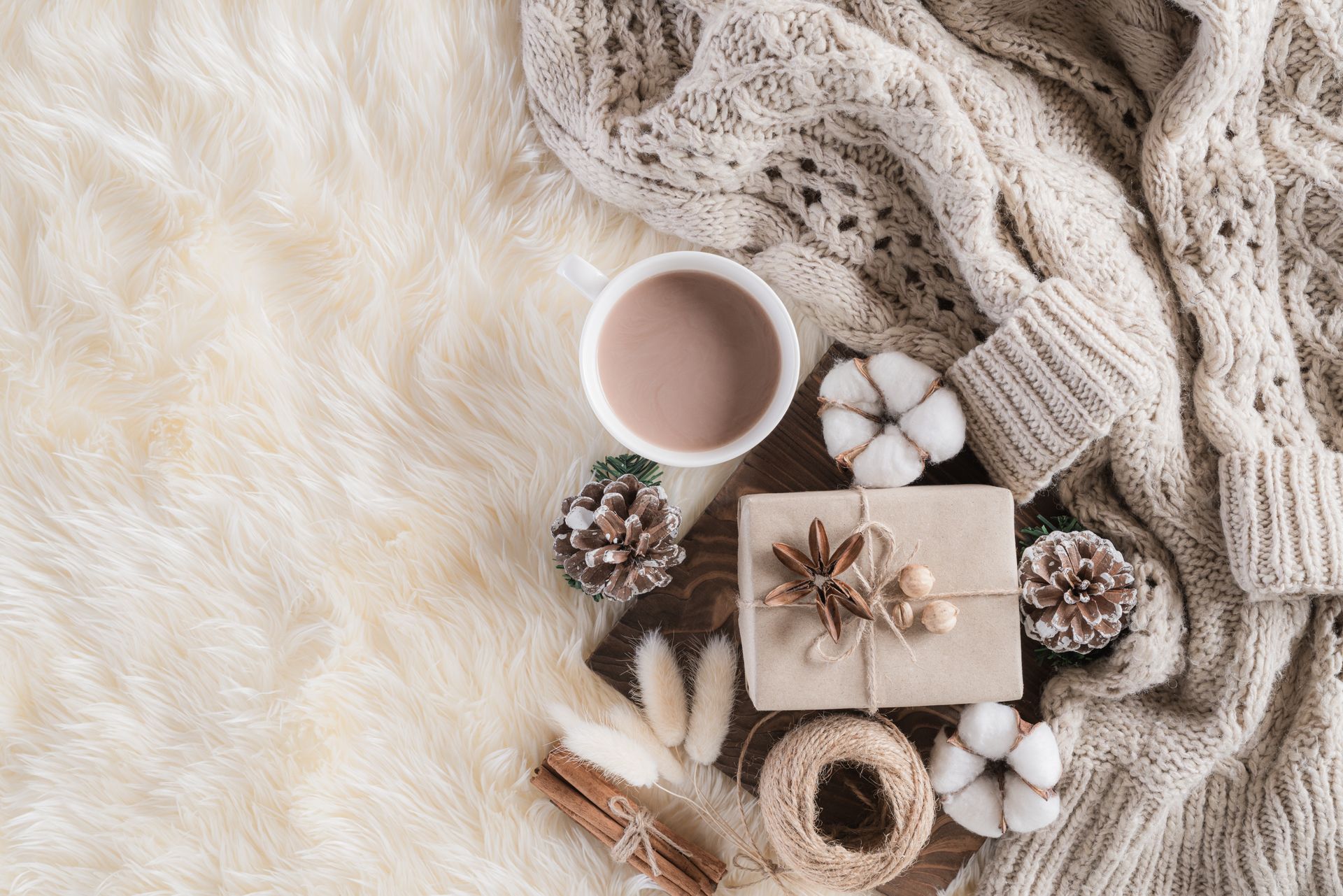 Cozy scene with a mug, gift, sweater, cotton stems, pinecones, and cinnamon on faux fur.