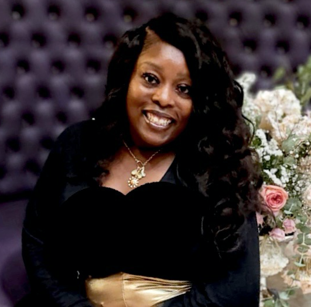 Woman with dark wavy hair smiles, wearing a black top and gold skirt, floral arrangement in the background.