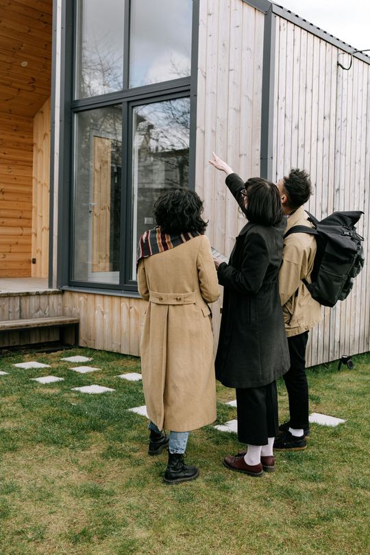 Three people stand outside a wooden building, looking up at the tall window.