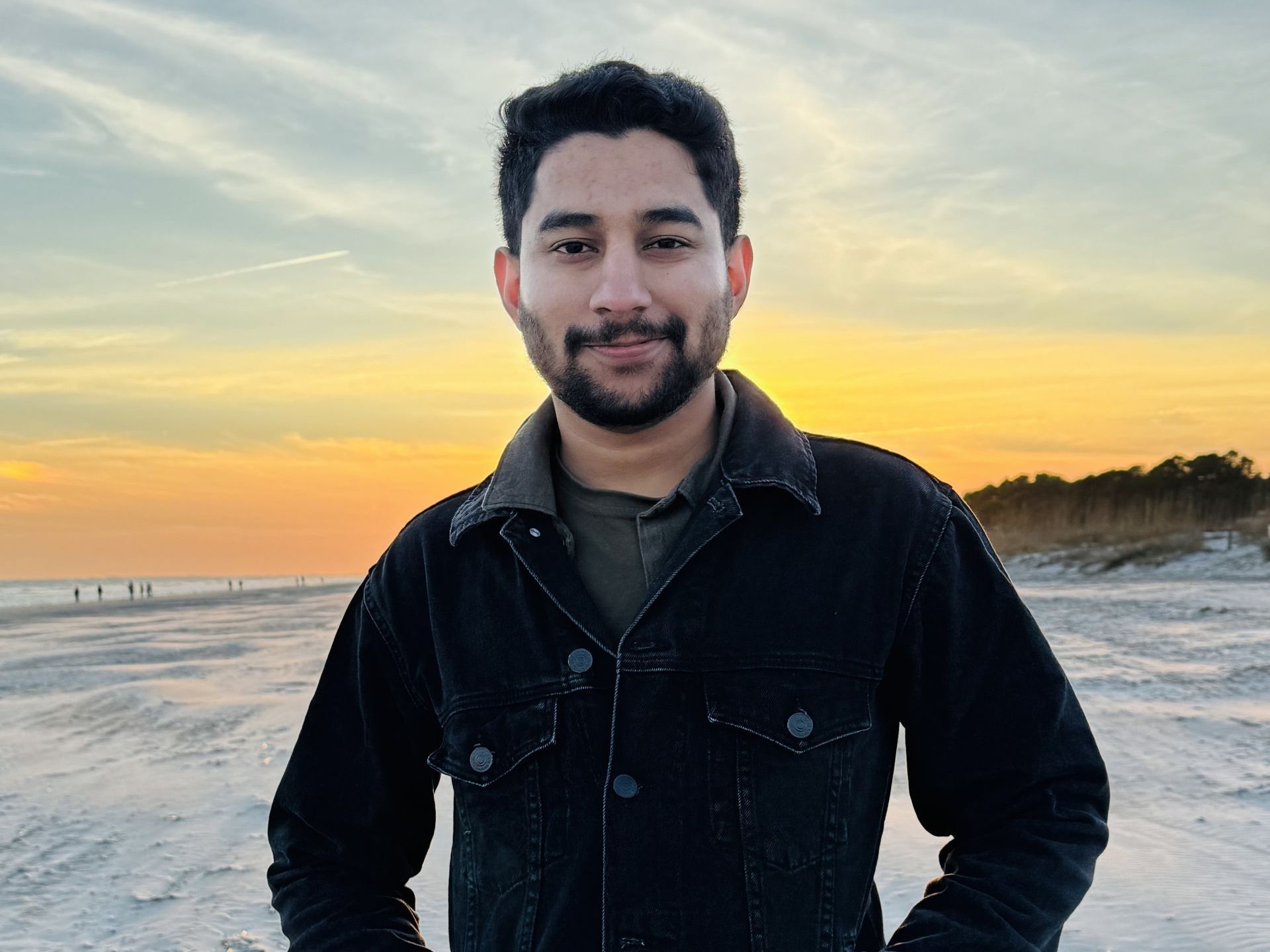 A man with a beard is standing on a beach at sunset.