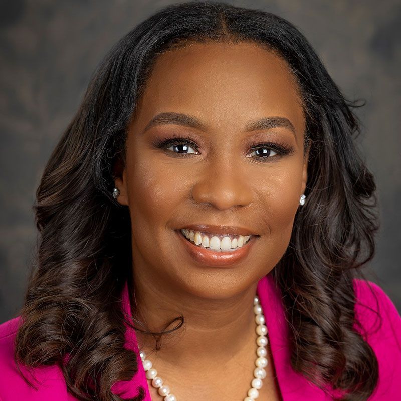 A woman wearing a pink jacket and pearls is smiling for the camera.