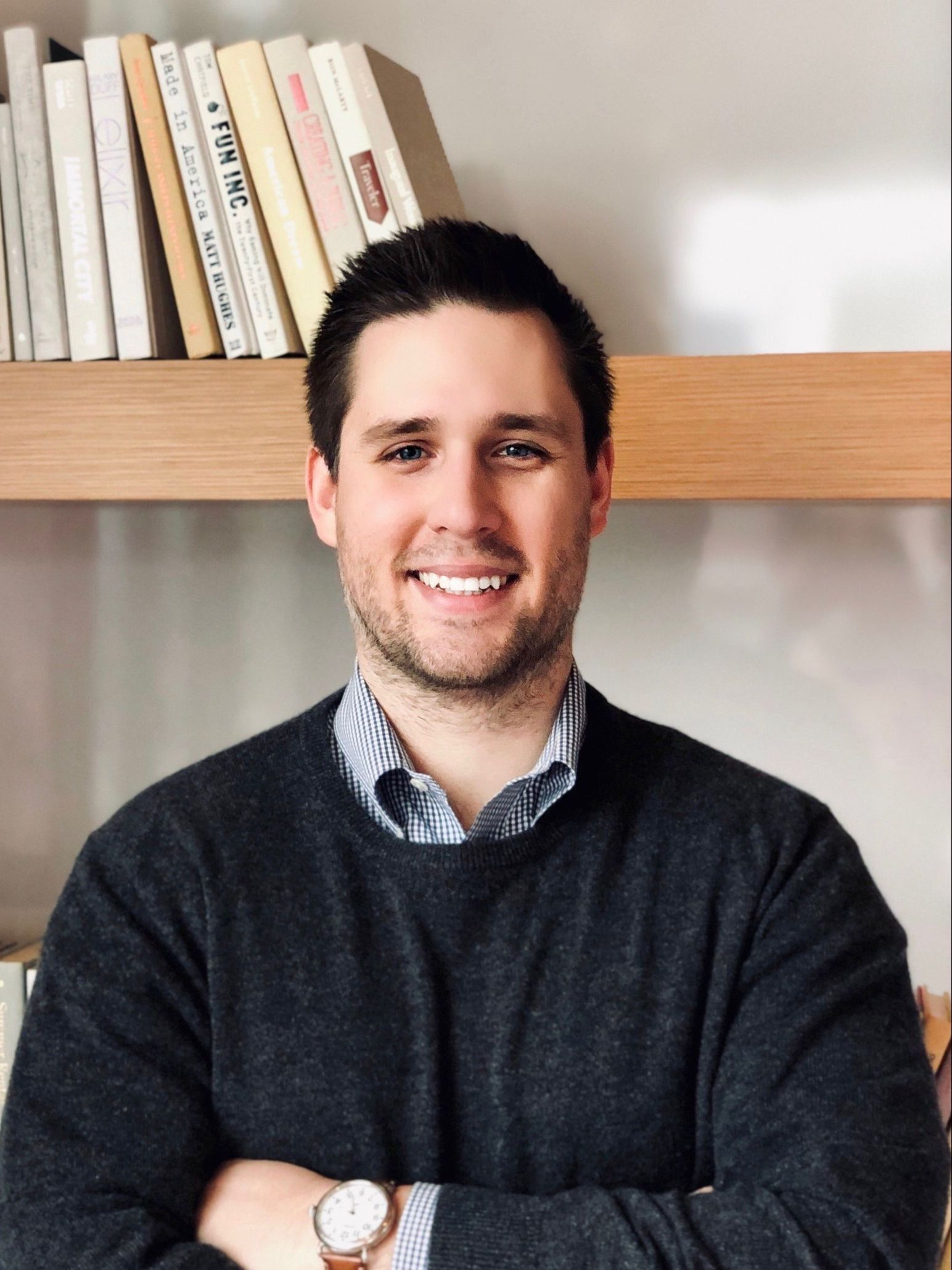 A man with his arms crossed is smiling in front of a bookshelf.