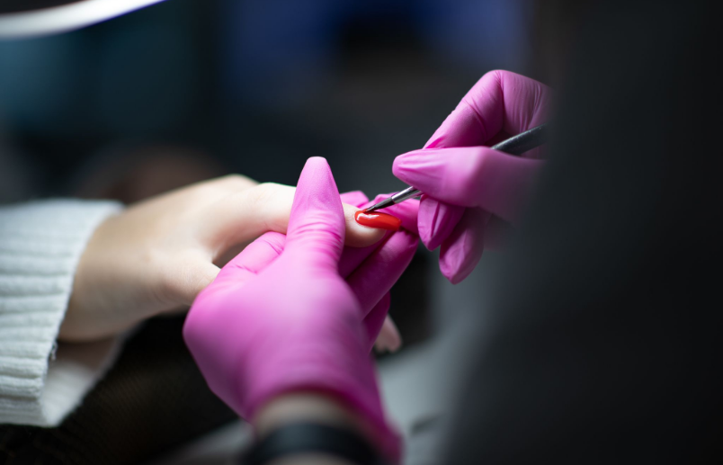 Eine Person, die sich die Nägel machen lässt; ihre Hände tragen rosa Handschuhe, ein Nagel wird rot lackiert.