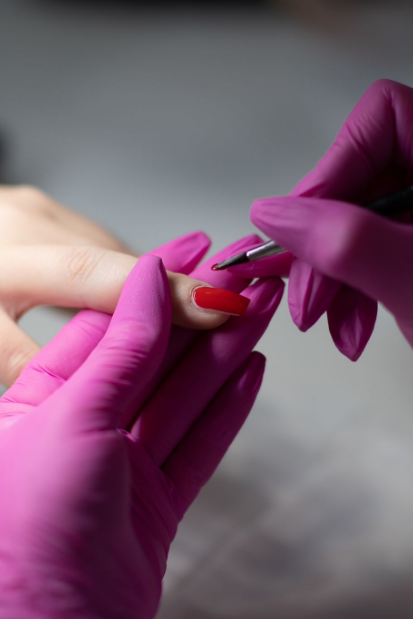 Hände in rosa Handschuhen tragen in einem Nagelstudio roten Nagellack auf einen Finger auf.