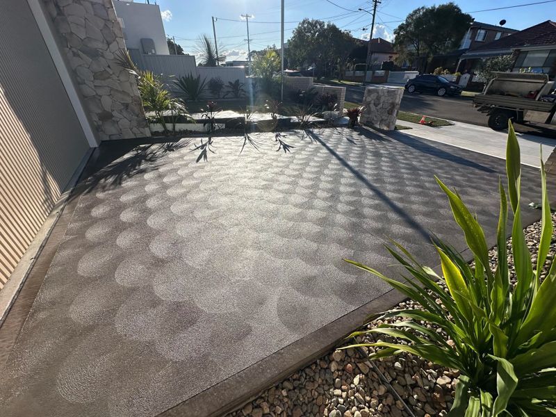 Sunny front patio with patterned concrete, potted plants, and a street view beyond the fence
