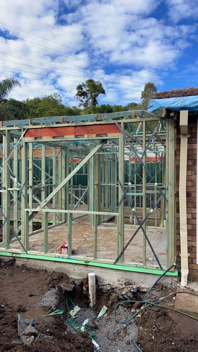 Partially built house with metal framing, green sheeting, and a muddy foundation under a cloudy sky