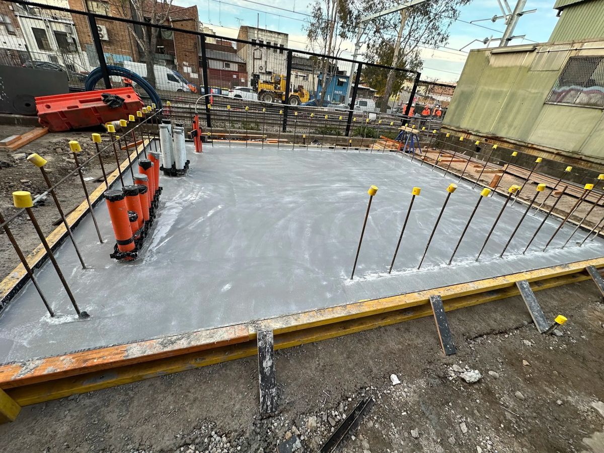 Concrete foundation under construction with rebar, orange barriers, and a fenced site backdrop