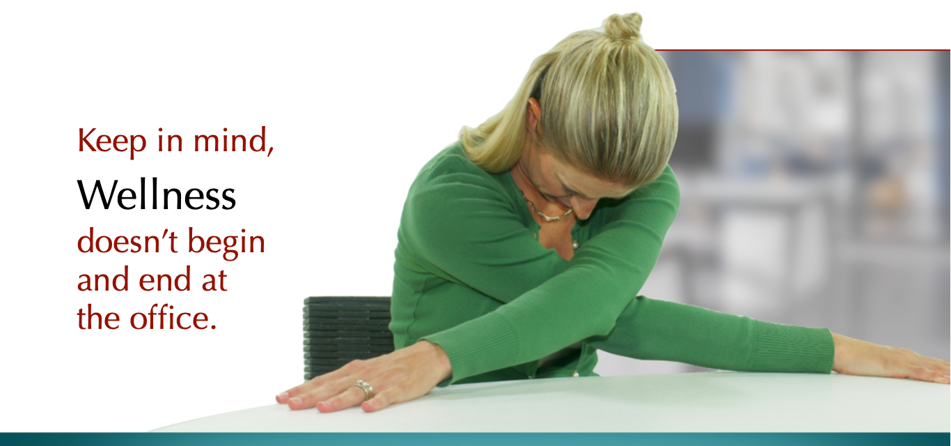 Keep in mind, Wellness doesn't begin and end at the office. A woman in a green sweater stretches at her desk.