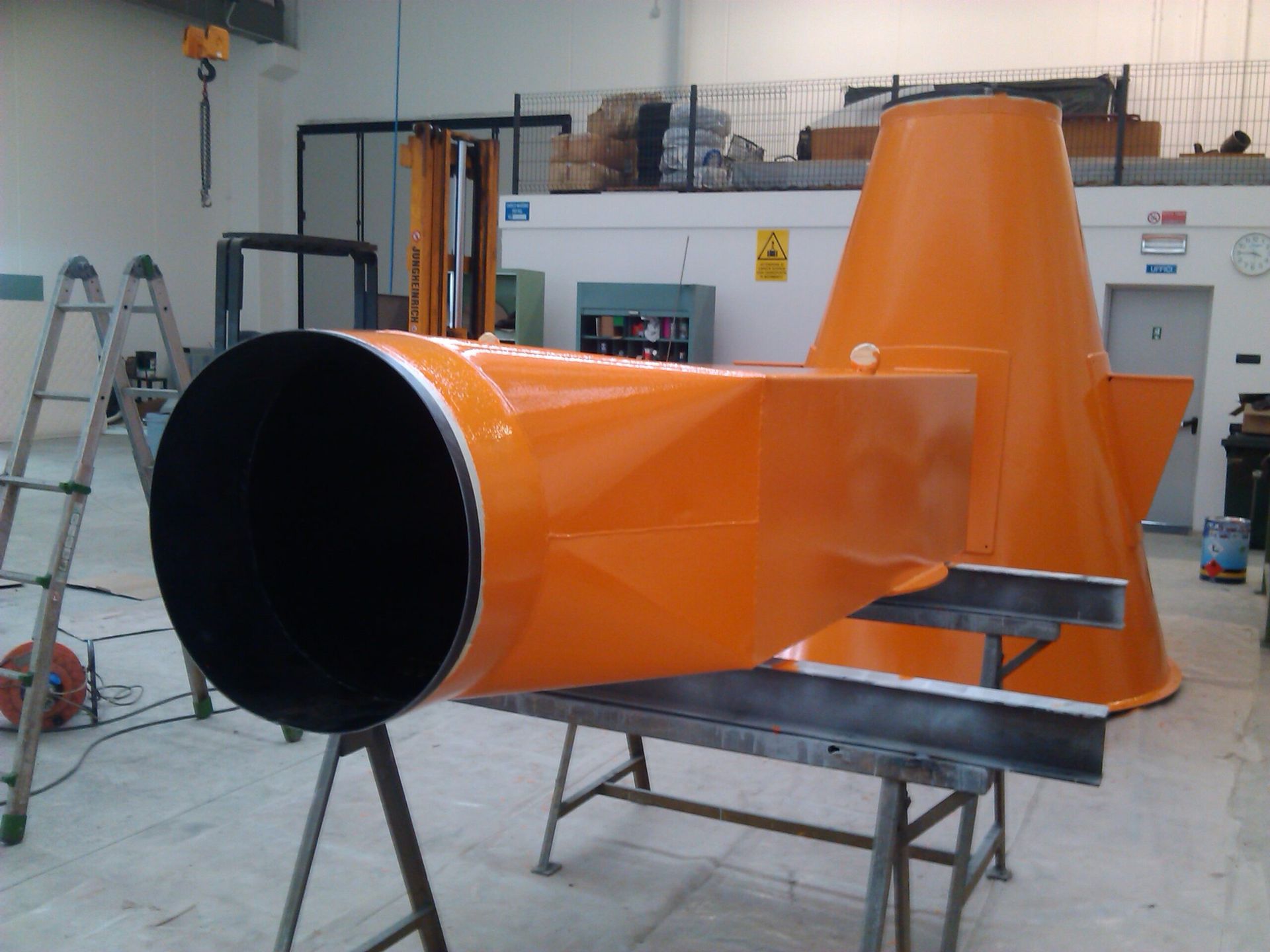 Condotto industriale arancione con apertura nera in un'officina, montato su supporti tra attrezzi e attrezzature.