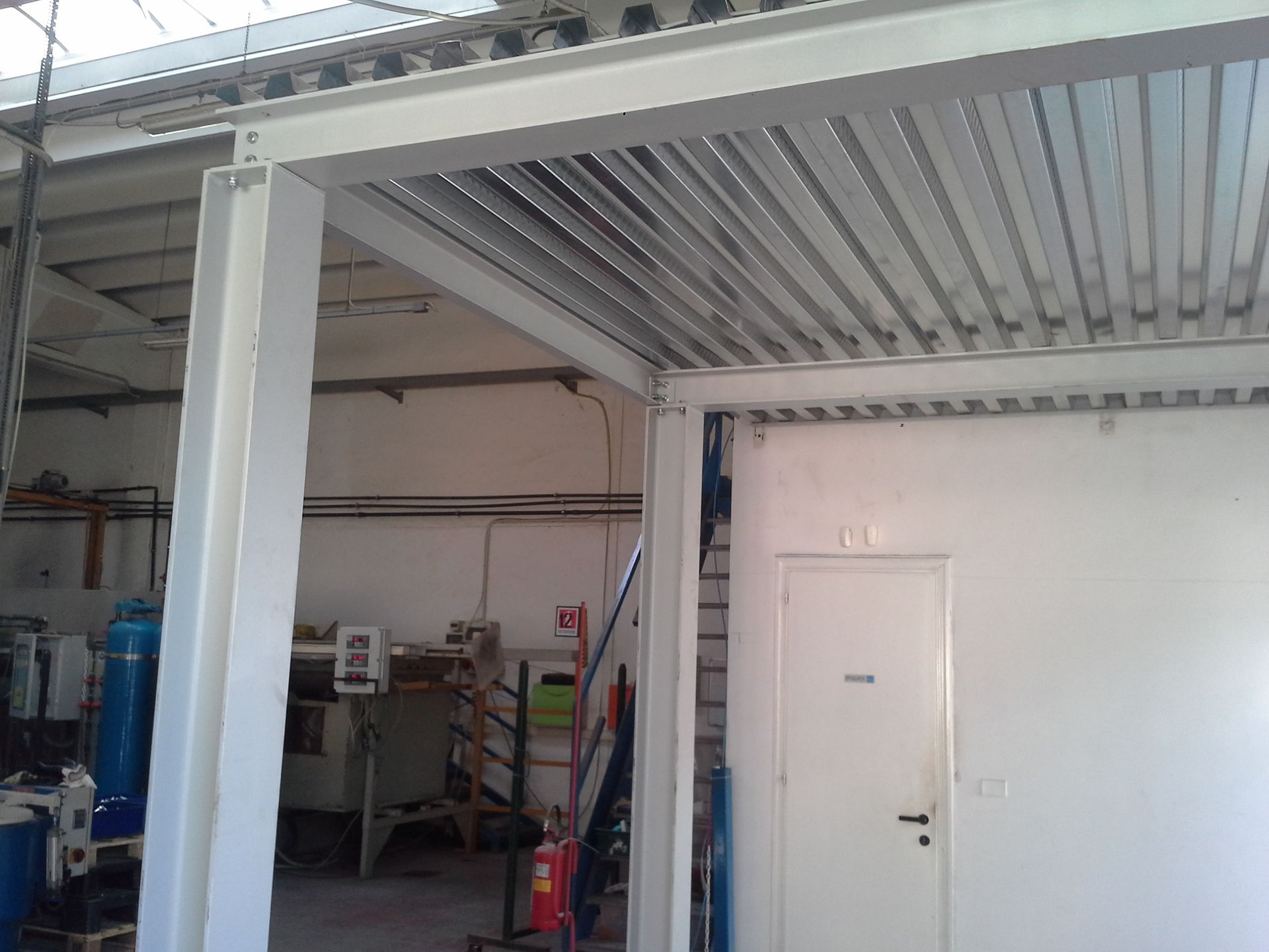 Interno industriale con porta bianca, soffitto in lamiera ondulata e attrezzature da officina sullo sfondo.