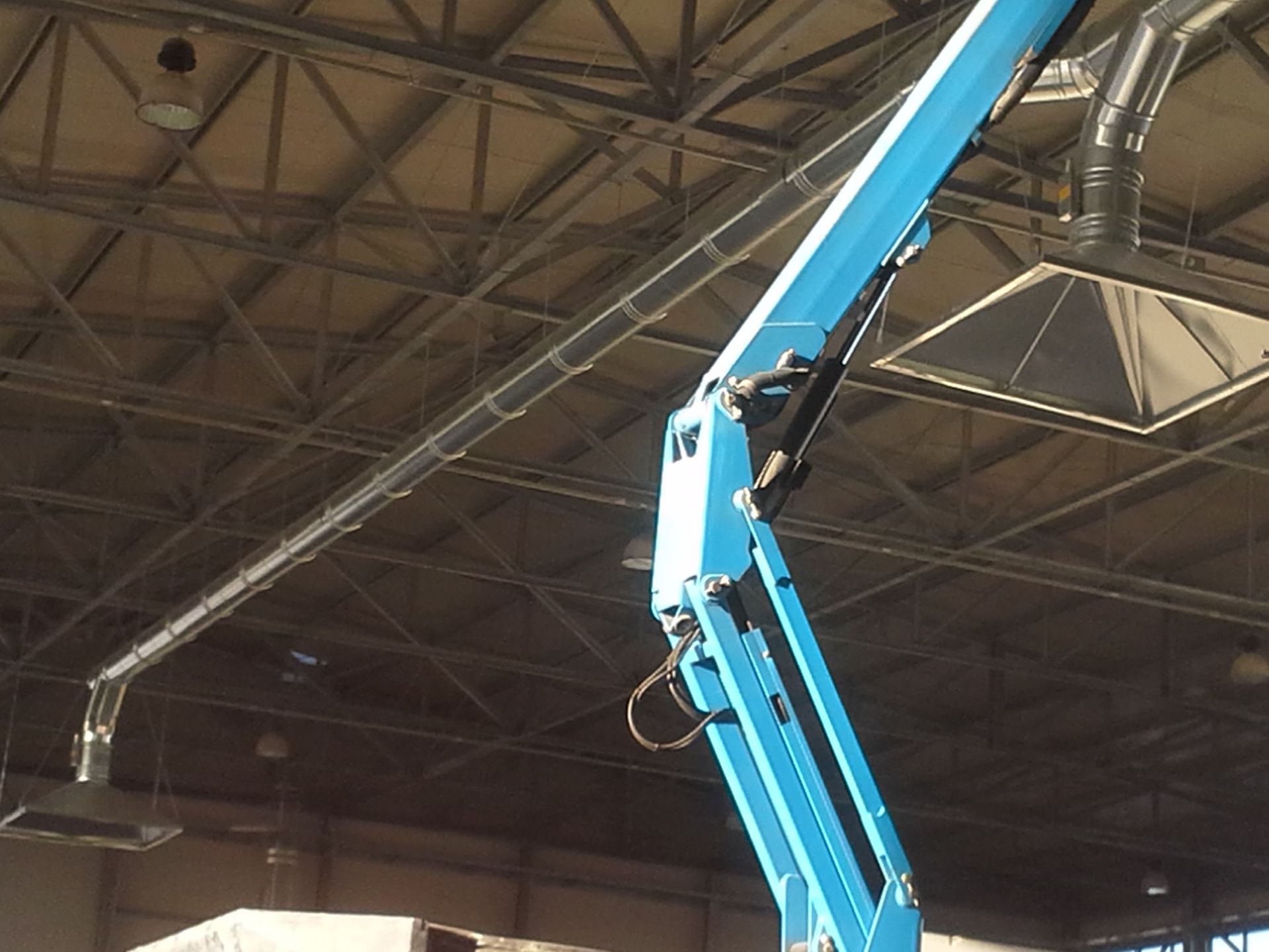 Piattaforma aerea blu sollevata all'interno di un magazzino industriale con soffitto a traliccio metallico.