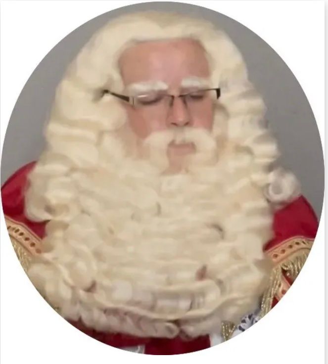 Persoon met een witte krullenpruik en bril, gekleed in een rood kostuum, tegen een grijze achtergrond.