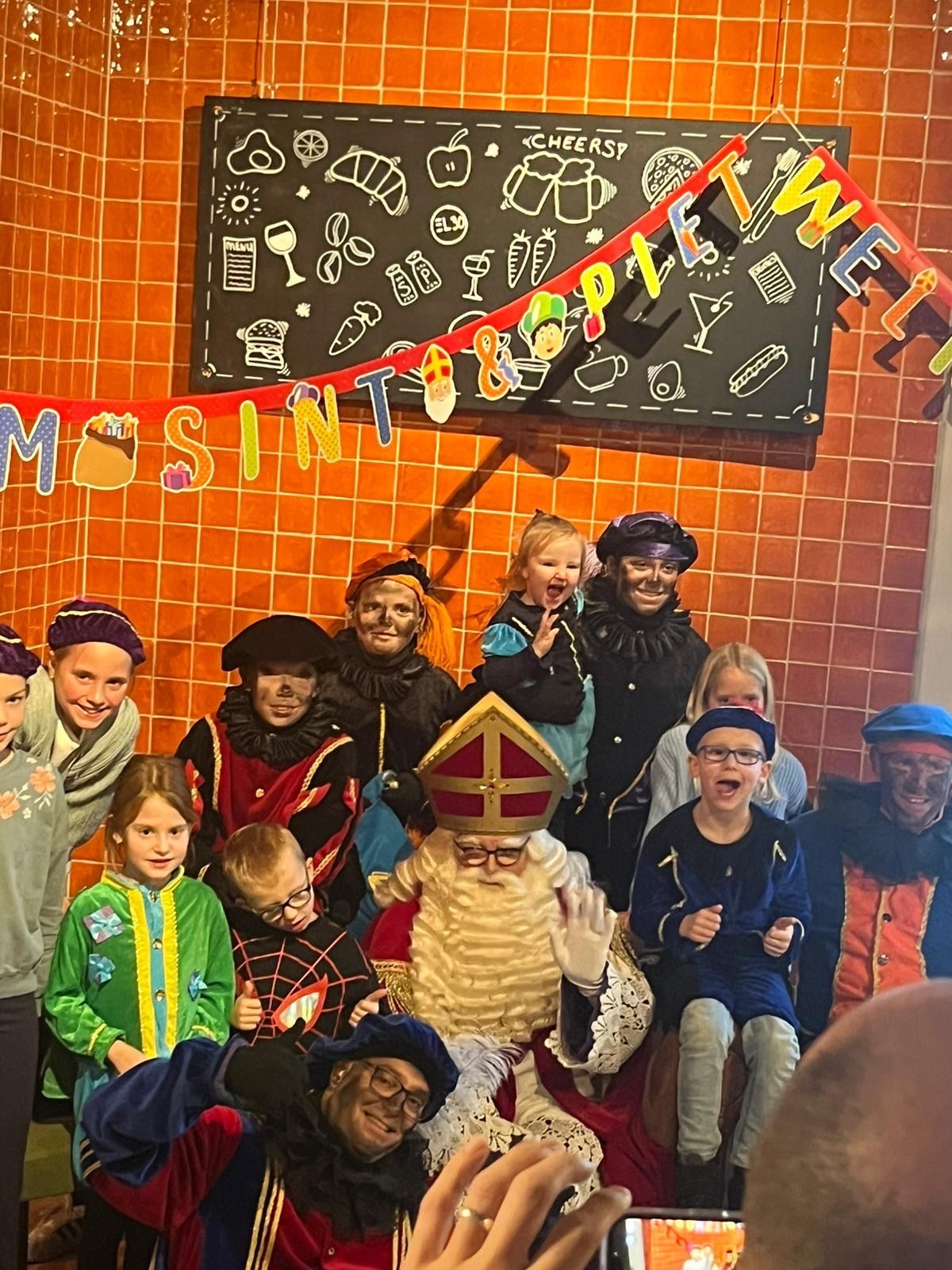Kinderen poseren met de kerstman voor een schoolbord en een oranje bakstenen muur, versierd met feestelijke kerstdecoraties.