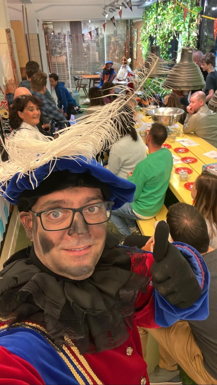 Persoon met blauwe feestmuts maakt een selfie op een levendig binnenfeest met kleurrijke versieringen en zittende gasten.