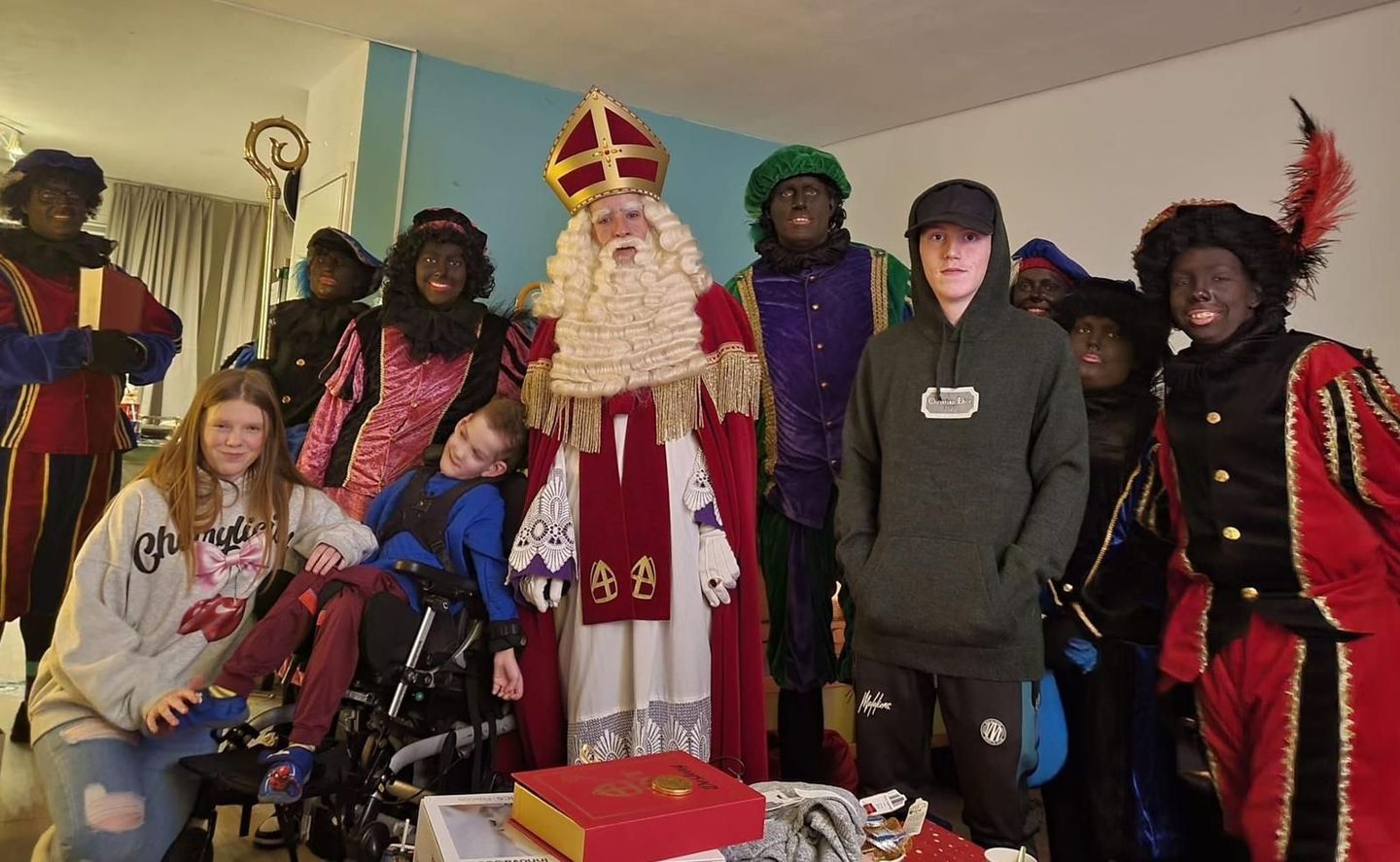 Mensen poseren binnen met Sint-Nicolaas in rode gewaden en een tafel vol geschenken ervoor.