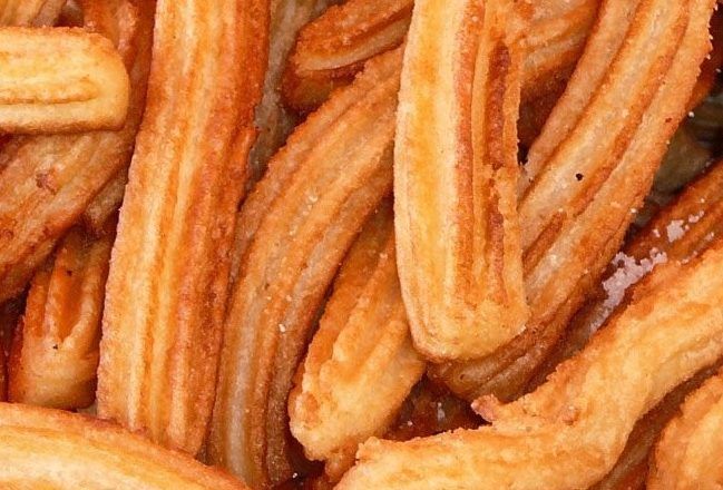 Close-up van goudbruine churros die op een hoop liggen en met suiker bestrooid zijn.