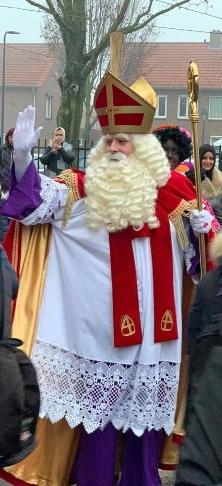 Sint Nicolaas in rode gewaden met een witte baard zwaait naar een feestelijke menigte in de openlucht.
