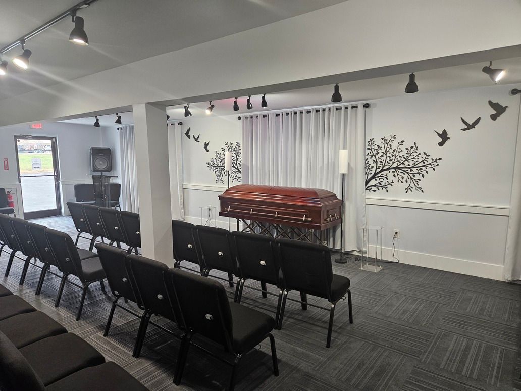 Small chapel with rows of black chairs, a wooden casket, and butterfly wall decor beneath track lighting