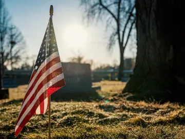American flag on a small pole in a sunlit grassy field, with bare trees and gravestones in the background