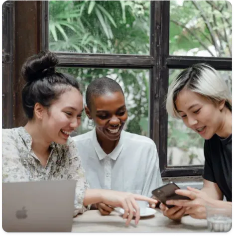 Three coworkers smiling and looking at a phone at a table with a laptop by a window