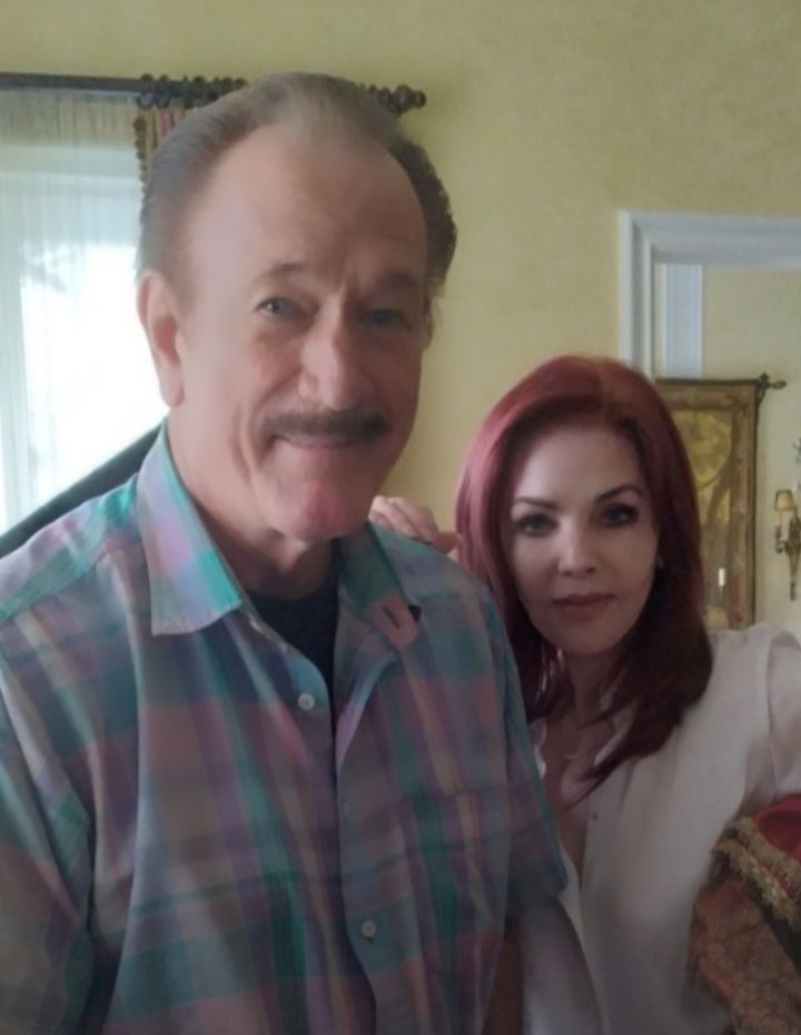A man with a mustache in a plaid shirt and a woman with red hair stand side-by-side in an indoor setting.