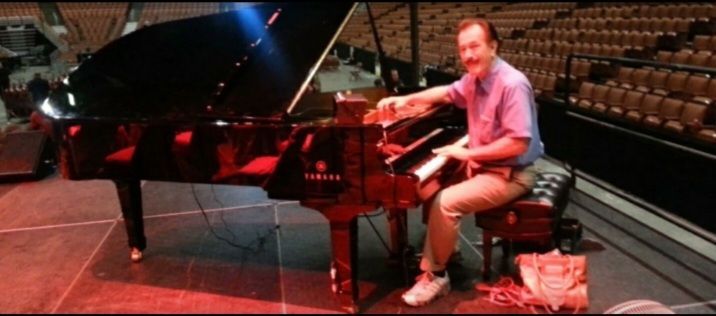 A person sits at a black Yamaha grand piano on a stage in an empty auditorium, smiling while playing the keys.