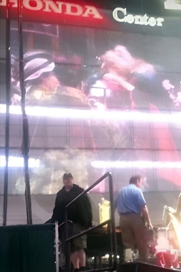 A large indoor screen at the Honda Center displays a close-up of a person, with two people standing in the foreground.