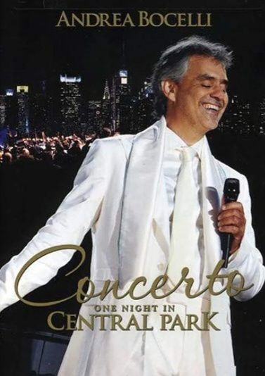 Andrea Bocelli in a white suit singing into a microphone on stage at night with a city skyline in the background.