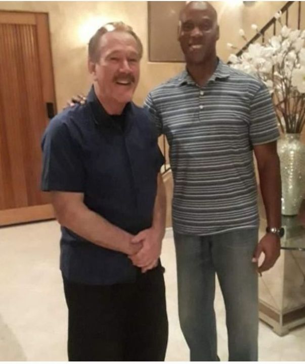 Two men smiling and posing for a photo indoors, standing in a hallway near a staircase and a decorative vase.