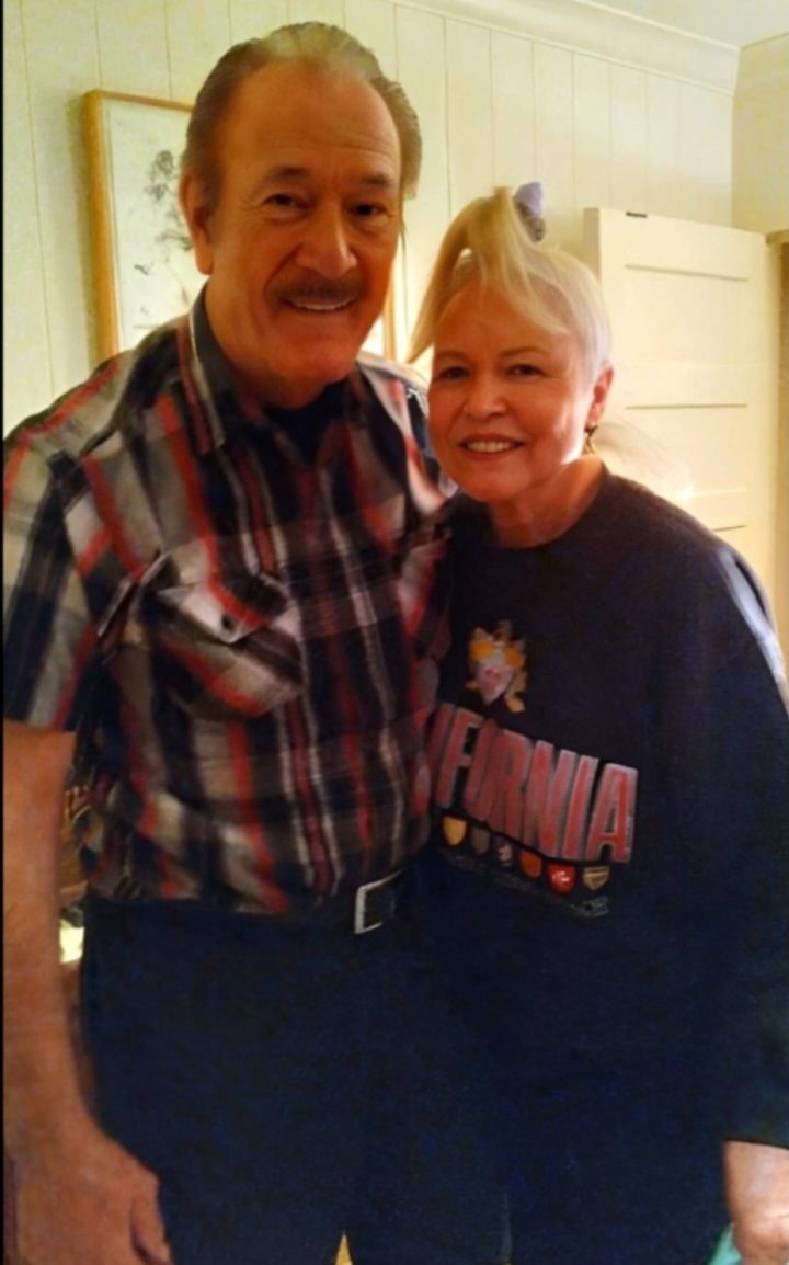 An older man in a plaid shirt and a woman in a black California sweatshirt smiling together indoors.