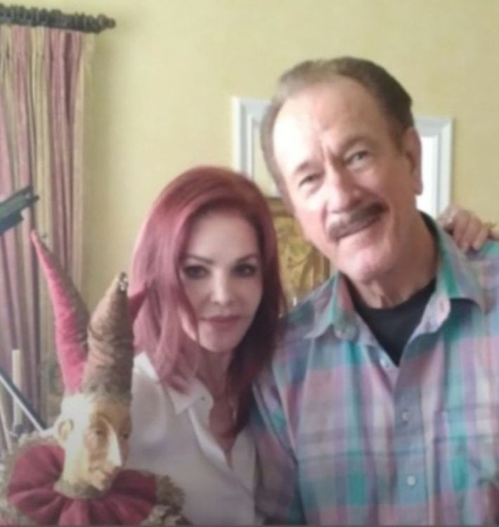 Priscilla Presley poses with Terry Gilliam next to a jester puppet, both smiling in an indoor setting.