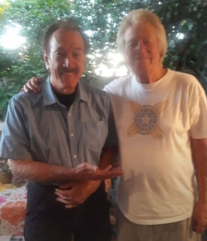 Two people stand outdoors with arms around each other, both smiling at the camera.