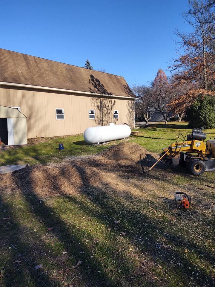 A large propane tank and a tree stump grinder near a tan building and a pile of wood chips on a sunny day.